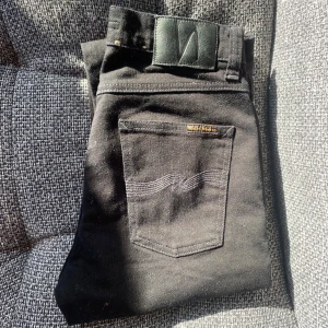 Svarta jeans från Nudie Jeans - Snygga svarta jeans från Nudie Jeans med klassisk femficksdesign och diskret broderad bakficka. Jeansen har en svart läderpatch i midjan och är tillverkade i mjukt denimtyg. Perfekta för dig som gillar stilrena och tidlösa byxor.