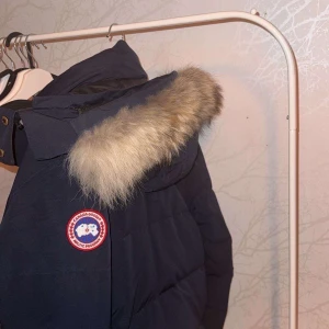 Mörkblå dunjacka från Canada Goose - Säljer en mörkblå dunjacka från Canada Goose med klassisk patch på ärmen. Jackan har huva med fluffig pälsdetalj, dragkedja och knappar framtill samt flera fickor. Perfekt för kalla vinterdagar och riktigt snygg streetstil.