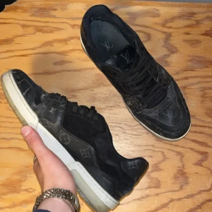 Louis vuttion trainers  - Snygga svarta sneakers från Louis Vuitton med klassiskt LV-monogram över hela skon. Skorna har svarta snören, vit och beige sula samt rund tå. Tillverkade i skinn med tydliga logodetaljer både på ovansidan och sulan. Perfekta för dig som vill sticka ut med lyxig streetstyle.