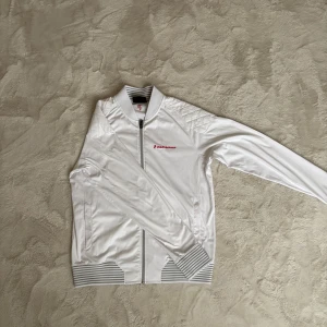 Vit Peak Performance M - Snygg vit tröja från Peak Performance med hel dragkedja och diskret logga på bröstet. Jackan har ribbade muddar med gråvita ränder och är tillverkad i 100% polyester. Perfekt för dig som gillar sportig och clean stil. Kan vara perfekt för dig som golfar eller en bra present 