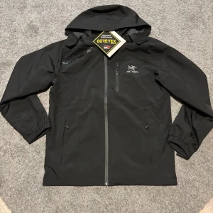 Svart Arc'teryx Gore-Tex vindjacka - Svart vindjacka från Arc'teryx med Gore-Tex-material som är vattentätt och andas. Jackan har huva, hel dragkedja framtill och diskret logga på bröstet. Perfekt för blåsiga och regniga dagar, med minimalistisk design och praktiska fickor.