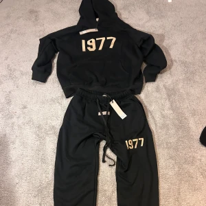 Svarta mjukisbyxor Essentials Fear of God - Svarta mjukisbyxor från Essentials Fear of God med beige '1977'-tryck på benet. Byxorna har elastisk midja med snörning och en diskret Essentials-logga framtill. Sköna och stilrena, perfekta för en avslappnad look.