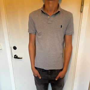 Grå pikétröja Polo Ralph Lauren - Klassisk grå pikétröja från Polo Ralph Lauren i slim fit-modell. Tröjan har korta ärmar, krage med knappar och den ikoniska broderade loggan på bröstet. Tillverkad i mjuk bomull som känns skön mot huden.