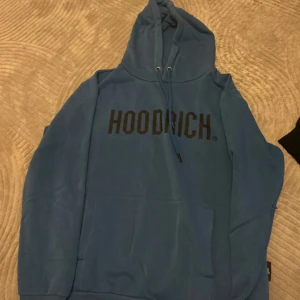 Blå hoodie från Hoodrich med tryck - Säljer en blå hoodie från Hoodrich i storlek medium. Tröjan har svart tryck framtill, huva med snörning och en stor magficka. Perfekt för dig som gillar streetwear och vill ha en skön och stilren look.