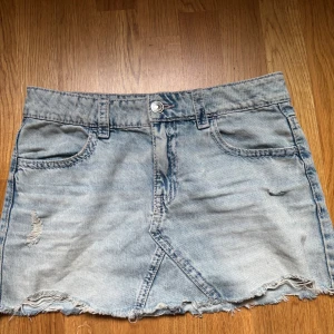 Ljusblå jeanskjol med slitna detaljer - Säljer en ljusblå jeanskjol med rå kant och slitna detaljer. Kjolen har klassiska fickor fram och bak, bälteshällor och snygga broderier på bakfickorna i rosa. Perfekt för dig som gillar en avslappnad och trendig stil.