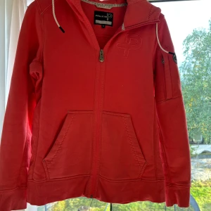 Rosa hoodie från Pelle P med huva - Snygg rosa hoodie från Pelle P med broderad logga på bröstet och patch på ärmen. Tröjan har huva med snörning, två stora fickor fram och dragkedja hela vägen. Tillverkad i mjuk bomull, perfekt för chill dagar. Passar dig som gillar sportig och avslappnad stil. Inga tecken på användning syns