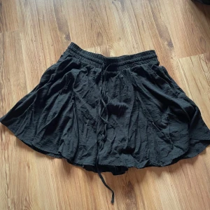 Svart byxkjol med snörning - Svart byxkjol med skön och luftig passform, tillverkad i ett mjukt och lätt material. Kjolen har resår i midjan och snörning framtill för extra justering. Perfekt för dig som gillar enkel och stilren design.