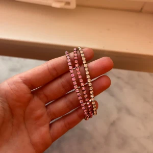 Guldiga armband med rosa, lila och silver stenar - Tre smala armband i guldton med gnistrande stenar, en med rosa stenar, en med lila stenar och en med silver stenar. Armbanden har en klassisk och stilren design.