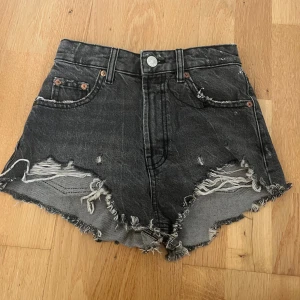 Svarta jeansshorts från Zara - Säljer ett par svarta, högmidjade jeansshorts från Zara i storlek 32. De har råa, fransiga kanter och slitna detaljer framtill för en edgy look. Klassisk femficksmodell med knapp och dragkedja. Perfekta för varma dagar och festivaler.