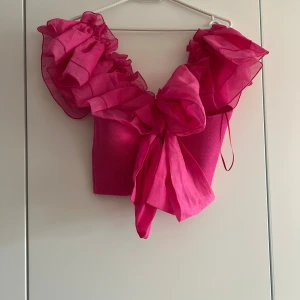 Rosa volangtopp med stor rosett - Superfin rosa topp med stora volangärmar i transparent material och en maffig rosett framtill. Toppen är croppad och har en riktigt statement-look, perfekt för dig som vill sticka ut. Materialet är en mix av polyester och ett lätt tyg som ger volym åt axlarna.