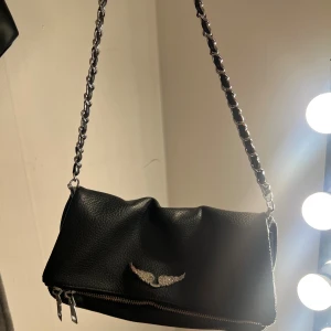 Svart axelväska lik Zadig väskorna - Snygg svart axelväska likt den självaste Zadig väskan  i skinnimitation med silverfärgad kedjerem och coolt emblem med glittrande vingar framtill. Väskan har dragkedja både fram och bak samt en extra ficka på baksidan. Ett längre band tillkommer dessutom! Kom privat för fler frågor💕