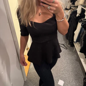 Svart peplumtop med fyrkantig ringning - Säljer en svart peplumtop med snygg fyrkantig ringning och halvlånga ärmar. Toppen har en figurnära passform upptill och ett utställd volang nedtill.