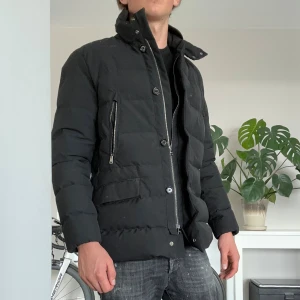 Svart dunjacka från Allegri - Snygg svart dunjacka från Allegri med hög krage, dragkedja och knappar framtill. Jackan har flera fickor med både dragkedja och lock, samt quiltad design som ger en clean och modern look. Perfekt för kalla dagar när du vill hålla stilen.