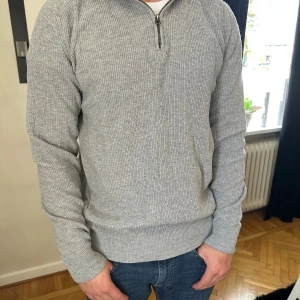Grå stickad half zip-tröja - Säljer en grå stickad tröja med half zip-dragkedja i halsen. Tröjan har lång ärm och ribbade muddar vid ärmslut och nederkant. Perfekt för dig som gillar en enkel och clean stil med lite sportig touch.