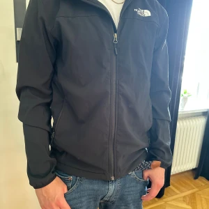 Svart vindjacka från The North Face - Svart vindjacka från The North Face med dragkedja framtill och två sidofickor med dragkedja. Jackan har en stilren design med logga på bröstet och ryggen. Perfekt för blåsiga dagar och enkel att matcha med jeans eller andra plagg. Kan gå ner till 400 vid snabb affär