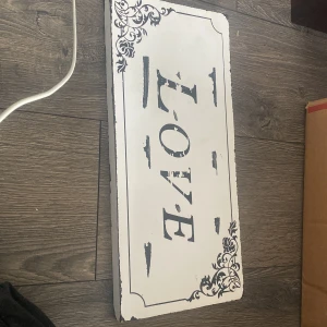 Tavla - Det här är ingen bok, utan en dekorativ skylt med texten 'LOVE' i vintage-stil och blommiga hörndekorationer. Perfekt för att skapa en mysig och inspirerande känsla i ditt rum eller på väggen.