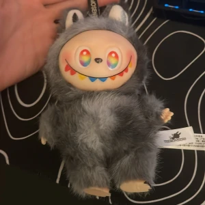 TheMonsters Plush Toy - Unik TheMonsters mjukisdocka med grå päls, färgglad leende och regnbågsögon. Dockan är i gott skick utan synliga defekter, perfekt för samlare eller som present. Märkt med originaletikett. Man kan diskutera priset 