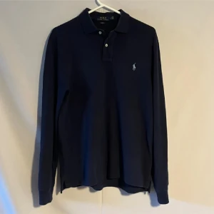 Mörkblå långärmad piké från Polo Ralph Lauren - Snygg mörkblå långärmad pikétröja från Polo Ralph Lauren med klassisk krage, knäppning med två knappar och broderad logga på bröstet. Tillverkad i mjuk bomull som känns skön mot huden. Perfekt för dig som gillar stilren och tidlös design.