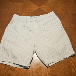 Randiga vita shorts från Kappahl - Fräscha vita shorts från Kappahl i storlek S med tunna vertikala ränder. De har elastisk midja med snörning och en bakficka. Perfekta för varma dagar och chill häng. Materialet känns lätt och skönt mot huden.