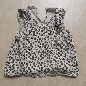Topp - Säljer en söt ärmlös topp från H&M i storlek 68. Toppen är vit med leopardmönster i brunt och svart, har volangdetaljer vid axlarna och knäppning bak i nacken. Perfekt för dig som gillar djurmönster och vill sticka ut lite extra.
