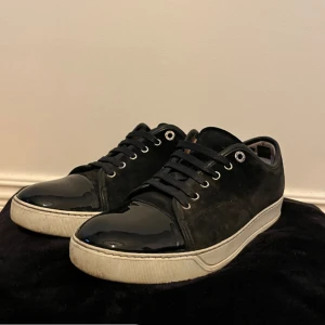Svarta sneakers från Lanvin i mocka - Snygga svarta sneakers från Lanvin med ovandel i mocka och lackad tå. Skorna har vita sulor, svarta skosnören och rund tå. Perfekta för dig som gillar en stilren och lyxig look med en twist av glans.