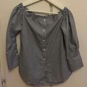 Randig offshoulder skjorta R&F S - Snygg randig skjorta från R&F i blått och vitt. Offshoulder-modell med långa ärmar och breda manschetter. Knappar längs ryggen ger en unik twist. Perfekt för dig som gillar trendiga detaljer och relaxed vibe.