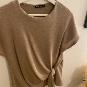 Beige stickad topp med knyt från Zara - Säljer en beige stickad topp från Zara med korta ärmar och rund halsringning. Toppen har en snygg knytdetalj i sidan. 