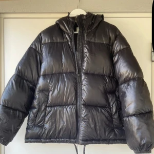 Svart dunjacka H&M - Säljer en svart pufferjacka i storlek S med huva och dragkedja framtill. Jackan har en glansig finish, två sidofickor och snörning nedtill. Perfekt för kalla dagar då den är tjock och håller värmen bra. Jackan är storlek S men lite större i storleken så skulle säga att den passer en S/M. Väldigt sparsamt använt så i mycket bra skick🥲