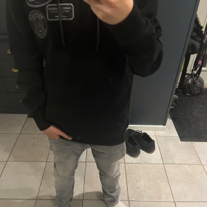 Svart hoodie från Mercier med patchar - Svart hoodie från Mercier med flera snygga broderade patchar på bröstet. Tröjan har huva med snörning och långa ärmar. Perfekt för dig som gillar streetwear och vill ha en stilren men ändå unik look. Säljer pga att jag inte tycker att den är så snygg.