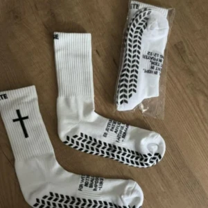 greppstrumpor kristna - Vita sportstrumpor från Rise Athlete med svart kors på skaftet och textcitat under foten. Strumporna har ribbad resår och svart mönster under sulan för extra grepp. Perfekta för dig som vill ha både stil och budskap i gymmet eller på planen. Helt oanvända (har flera par)