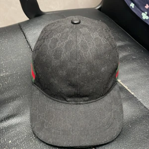 Svart keps från Gucci med monogram - Säljer en svart keps från Gucci med diskret GG-monogram över hela ytan. Kepsen har klassisk form, justerbart spänne bak i svart skinn och gröna/röda detaljer på sidorna. Materialet är bomull och detaljer i skinn. Perfekt för dig som gillar exklusiva accessoarer.
