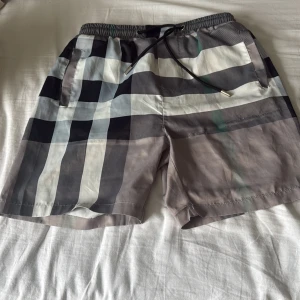Grå burrberry shorts - Snygga Burberry shorts med klassiskt rutigt mönster i svart, grått och vitt. De har elastisk midja med snörning och sidofickor, samt en bakficka. Perfekta för sommaren och har en loose fit. Materialet känns lätt och svalt, perfekt för varma dagar.