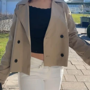 Beige trenchcoat från Vila - Snyggtrenchcoat från Vila i beige med dubbelknäppning och stora svarta knappar. Jackan har klassisk krage och är perfekt för dig som vill ha en trendig och stilren look.
