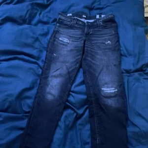 Slim svarta jeans Jack & Jones 164 - Snygga svarta slim fit jeans från Jack & Jones, modell Glenn. Jeansen har slitna detaljer och hål vid knäna för en cool look. Klassisk femficksmodell med dragkedja och knapp. Perfekta för dig som gillar en smalare passform och streetstil.