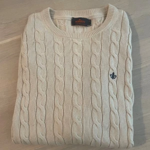 Beige kabelstickad tröja från Morris - Kabelstickad beige tröja från Morris i 100% pima bomull. Klassisk rund halsringning, ribbade muddar och diskret broderad logga på bröstet. Perfekt för dig som gillar stilren och tidlös design. Kan ha små tecken på användning men är i väldigt bra skick.
