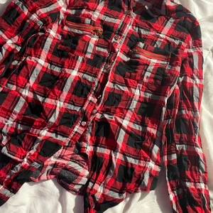 Röd och svart rutig skjorta Foresta Bella - Rutig skjorta från Foresta Bella i rött, svart och vitt. Skjortan har långa ärmar, knappar framtill och två bröstfickor med coola dragkedjor. Perfekt för dig som gillar en avslappnad och trendig stil.