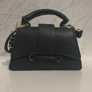 Svart handväska i skinn med axelrem - Stilren svart handväska i präglat skinn med både handtag och justerbar axelrem. Väskan har guldfärgade metalldetaljer och snygga ringar framtill. Perfekt storlek för det viktigaste och passar till många outfits.