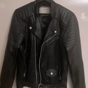 Svart skinnjacka från Marty & Gus - Snygg svart skinnjacka från Marty & Gus med coola detaljer som sned dragkedja, bälte i midjan och quiltade axlar. Jackan har flera fickor med dragkedja och tryckknappar, samt klassisk bikerstil. Perfekt för dig som vill ha en edgy look.