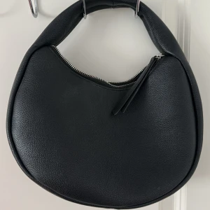 Svart axelväska i skinnlook - Stilren svart axelväska i rundad form med dragkedja upptill. Väskan har en mjuk struktur och är tillverkad i skinnimitation. Perfekt för dig som vill ha en enkel men trendig accessoar till vardagslooken.