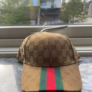 Gucci keps med GG-mönster och röd/grön rand - Snygg keps från Gucci i beige med klassiskt GG-monogram över hela kepsen. Framtill finns en bred röd och grön rand som ger en ikonisk look. Justerbar rem baktill med metallspänne där Gucci är ingraverat. Tillverkad i bomullsmix.