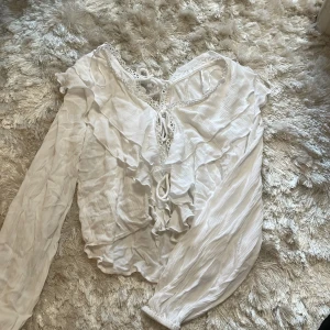 Vit blus med volanger och snörning - Supersöt vit blus med långa ärmar, volangdetaljer och snörning framtill. Blusen har v-ringning med spetskant och är gjord i ett lätt, krinklat tyg som ger en romantisk vibe. Perfekt för dig som gillar en feminin och trendig look.