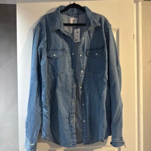 Ljusblå jeansskjorta från Lager 157 - Säljer en ljusblå jeansskjorta i storlek L. Skjortan har klassisk krage, två bröstfickor med knappar och långa ärmar. Tillverkad i jeansmaterial och stängs med tryckknappar framtill. Perfekt för en avslappnad och cool look.