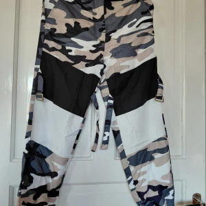 Cargopants med camomönster och snörning - Säljer två par cargopants med camomönster. Ena paret har beige, svart och vit camo med bältesdetaljer och snörning i midjan. Det andra paret har grå, svart, vit och rosa camo med resår och snörning. Perfekta för dig som vill sticka ut med streetstyle. Stl: S/36 Mitt pris: 100:- styck