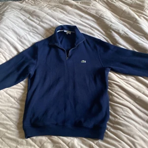 Half zip tröja från Lacoste - Snygg marinblå tröja från Lacoste med klassisk krokodillogga på bröstet. Tröjan har half zip-dragkedja, hög krage och långa ärmar. Perfekt för lager-på-lager och chill stil. Materialet är mjukt och känns riktigt skönt mot huden.