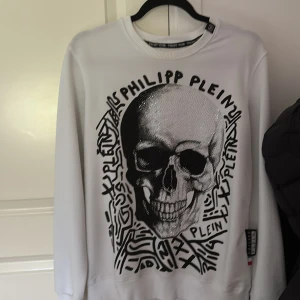 Vit sweatshirt från Philipp Plein - Säljer en vit sweatshirt från Philipp Plein med svart grafiskt tryck och logga framtill. Tröjan har rund hals, långa ärmar och en snygg PP-detalj i nacken. Mjuk och skön bomullskänsla, perfekt för dig som gillar streetstyle och statement-plagg.