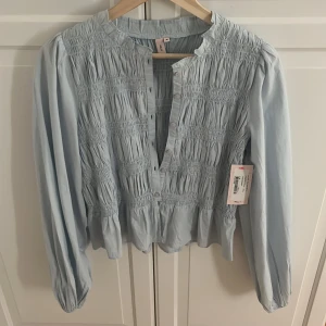 Ljusblå blus med smock från Nelly - Supersöt ljusblå blus från Nelly med smockad överdel och långa ärmar. Blusen har knappar framtill och en lätt volang vid nederkanten. Aldrig använd med prislapp kvar!