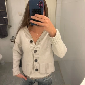 Beige stickad kofta med stora knappar - Superfin beige stickad kofta i kashmir och ull med bred ribbad kant och stora mörka knappar framtill. Koftan har v-ringning och långa ärmar, perfekt att slänga på sig över ett linne eller t-shirt. Snygg och enkel att styla till jeans eller kjol.