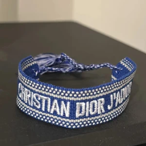 Blått armband från Christian Dior - Snyggt blått armband från Christian Dior med texten 'CHRISTIAN DIOR J'ADIOR' broderad i vitt. Armbandet är gjort i bomull och har justerbara knytband med tofsar i blått och vitt. Perfekt accessoar för att lyfta din outfit.