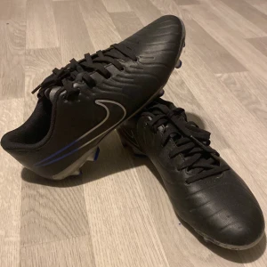 Svarta Nike fotbollsskor med blå detaljer - Säljer ett par svarta Nike fotbollsskor med blå detaljer och silverfärgad sula. Skorna har snörning och är tillverkade i syntetmaterial med klassisk Nike-logga på sidan. Perfekta för dig som vill ha stilrena och sportiga skor på planen.