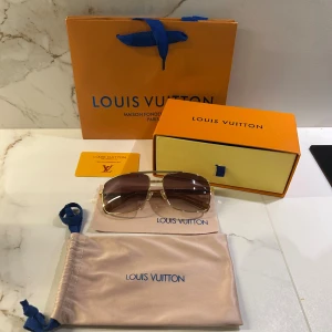 Louis Vuitton Attitude solglasögon guldbrun - Snygga Louis Vuitton solglasögon med fyrkantig form och guldfärgad metallbåge. Glasen är bruna med en snygg toning. Kommer med originalfodral, ask och påse. Perfekt accessoar för att lyfta din stil.